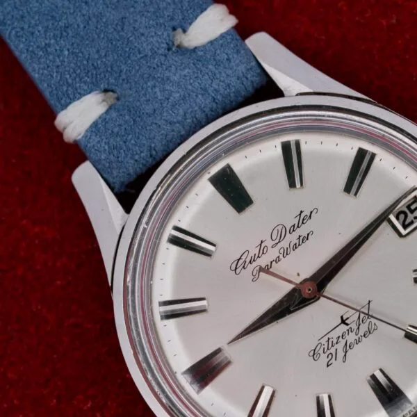 Close-up of a Citizen Jet Auto Dater Automatic Para Water wristwatch with silver dial, black hour markers, a date window displaying 25, and a blue suede strap against a vibrant red background.