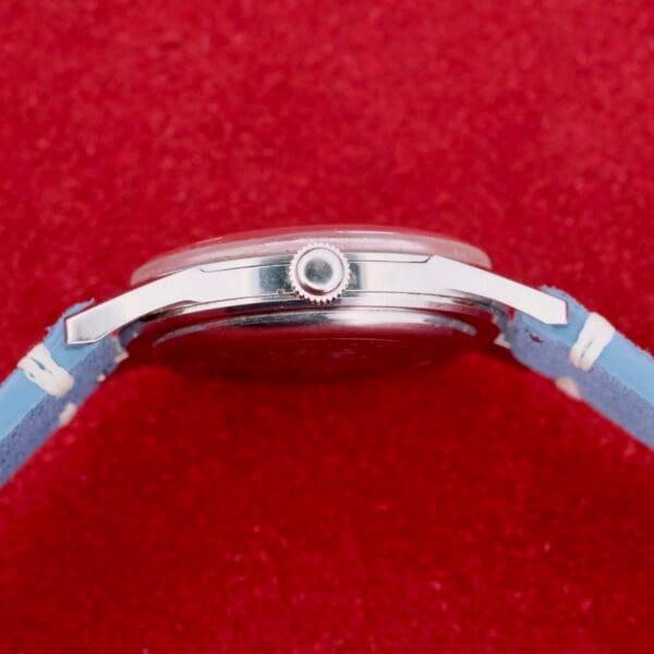 Side view of a Citizen Jet Auto Dater Automatic Para Water watch with a blue leather strap on a red textured background. The silver case features a visible crown on the right side, highlighting timeless elegance and precision.