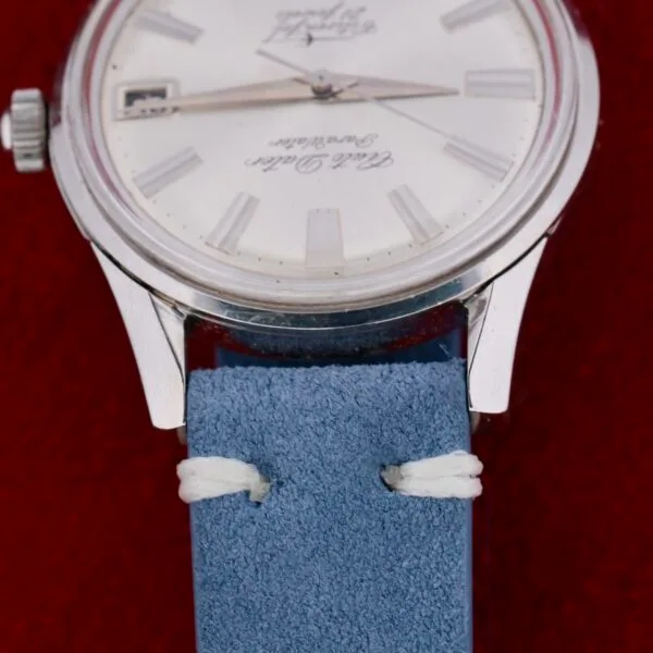 Close-up of a vintage Citizen Jet Auto Dater Automatic Para Water wristwatch with a silver case, white dial, and date display. Its secured with a light blue suede strap featuring white stitching against a deep red background.