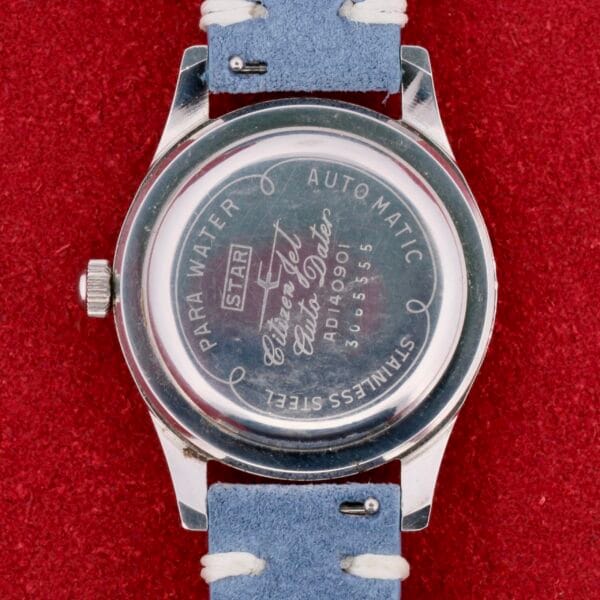 A wristwatch sits on a red surface, showing Star, Para Water, Automatic, and model numbers. The Citizen Jet Auto Dater Automatic Para Water has a silver casing with a light blue leather strap, crafted from stainless steel for timeless appeal.