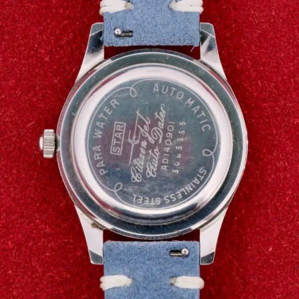A wristwatch sits on a red surface, showing Star, Para Water, Automatic, and model numbers. The Citizen Jet Auto Dater Automatic Para Water has a silver casing with a light blue leather strap, crafted from stainless steel for timeless appeal.