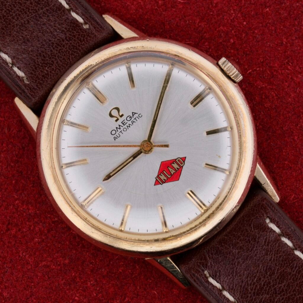 A vintage Omega Automatic Cal. 551 for Inland Company wristwatch with a brown leather strap sits on a red textured surface, showcasing a silver face with gold hour markers, hands, and the iconic Omega logo along with the word Inland in red.