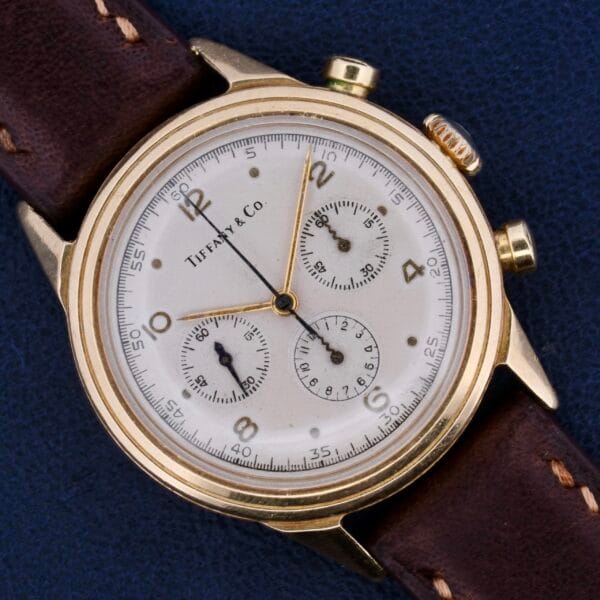 A Vintage Movado For Tiffany & Co 14k Gold Chronograph M95 watch, featuring a white face with three subdials and a brown leather strap, is elegantly showcased on a blue surface.