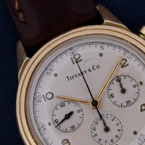 A close-up of a Vintage Movado for Tiffany & Co 14k Gold Chronograph M95, featuring a white face with gold casing and three subdials, gold hour and minute hands, Arabic numerals marking the hours on a unique Odd Dot Dial against a dark background.