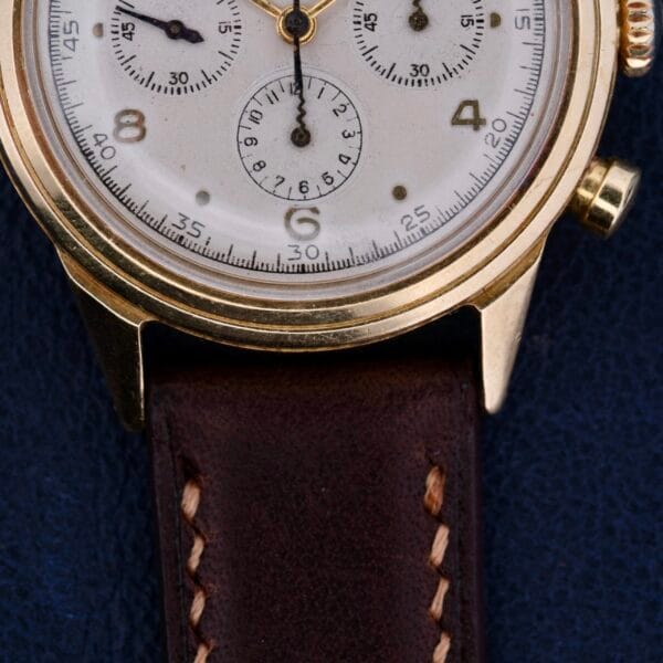 Close-up of a Vintage Movado For Tiffany & Co 14k Gold Chronograph M95 wristwatch with an odd dot dial, white face, and brown leather strap. It rests on a dark blue surface, exuding the timeless sophistication typical of Tiffanys esteemed collections.