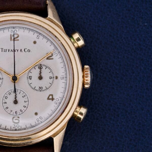 Close-up of a Vintage Movado for Tiffany & Co 14k Gold Chronograph M95 - Odd Dot Dial. The exquisite timepiece features three subdials, gold-tone hands, and a leather strap set against a blue textured background.
