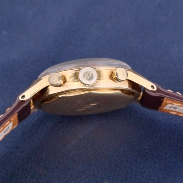 Side view of the Vintage Movado For Tiffany & Co 14k Gold Chronograph M95 with a round case, chronograph buttons, and textured crown. It’s paired with a brown leather strap on a dark blue surface.