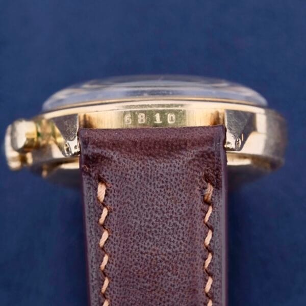 A close-up of the Vintage Movado For Tiffany & Co 14k Gold Chronograph M95 - Odd Dot Dial wristwatch with a brown leather strap, reminiscent of early Movado designs. The side shows 6810 on a deep blue background.