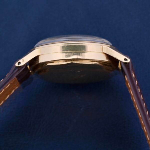 A side view showcases the elegance of the Vintage Movado for Tiffany & Co 14k Gold Chronograph M95, featuring a slim profile and gold casing, with a brown leather strap against a dark blue surface.