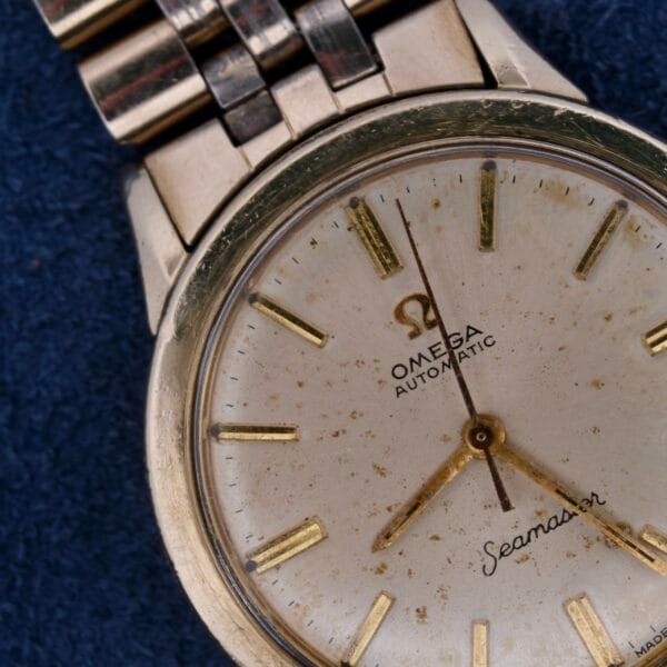 Close-up of a vintage Omega Seamaster Automatic Cal. 550 wristwatch with a metallic band. The dial shows an aged white background, gold hour markers, and hands with signs of wear and patina, along with the brand name and logo clearly visible.