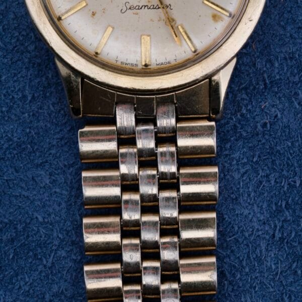 Close-up of a vintage Omega Seamaster Automatic Cal. 550 watch on a textured blue surface, featuring Seamaster on its round face, minimalistic hour markers, and a metal link bracelet.