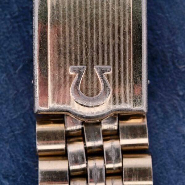 Close-up of a gold Omega Seamaster Automatic Cal. 550 watch clasp with a horseshoe logo. The brushed metal surface and segmented band contrast with the polished edges and logo against the blue background.