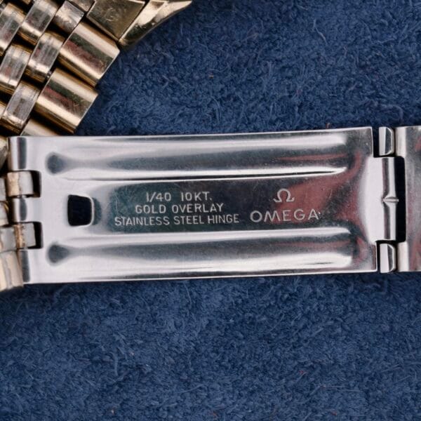 A close-up of the Omega Seamaster Automatic Cal. 550 watch band clasp on a textured blue surface features an engraving: 1/40 10KT GOLD OVERLAY STAINLESS STEEL HINGE beside the Omega logo. The bracelets gold and silver links exude timeless luxury.