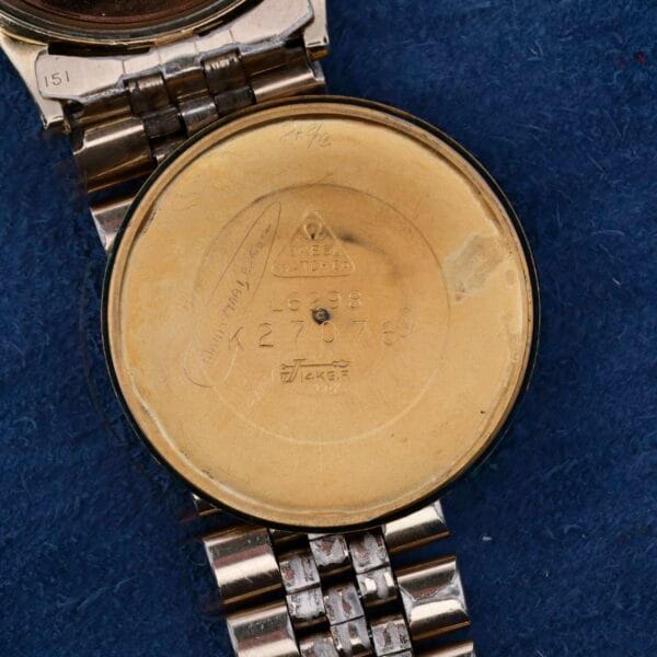 A close-up of an Omega Seamaster Automatic Cal. 550 watch case back reveals engraved text and numbers, with its automatic design highlighted by a metal strap against a textured blue fabric backdrop.