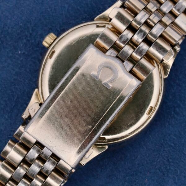 A close-up of a gold Omega Seamaster Automatic Cal. 550 wristwatch with a metal link band displays the iconic logo on the clasp, elegantly resting on a dark blue fabric background.