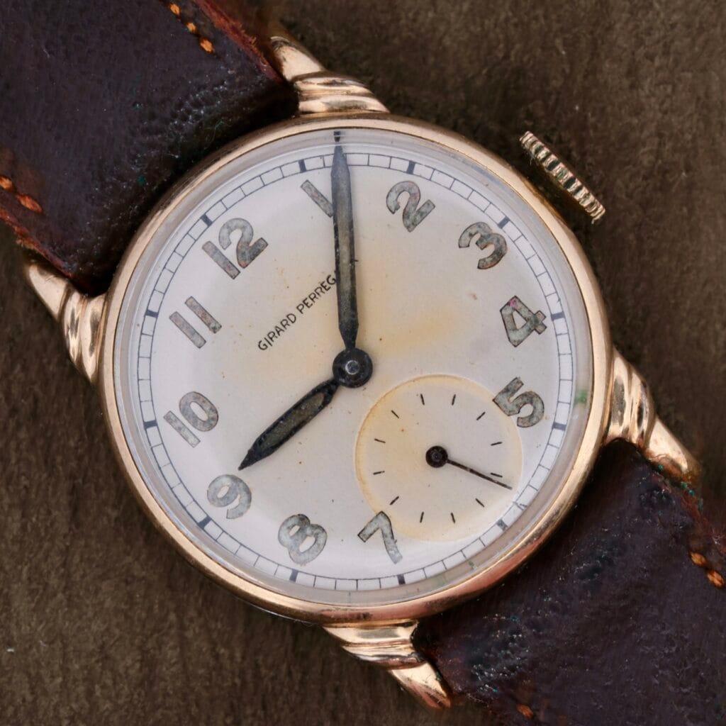 The Girard Perregaux Military Dial Small Seconds is a vintage wristwatch with a white face, black numerals, and gold casing. It features black hour and minute hands, a small seconds subdial at 6 oclock, and a dark worn leather strap.