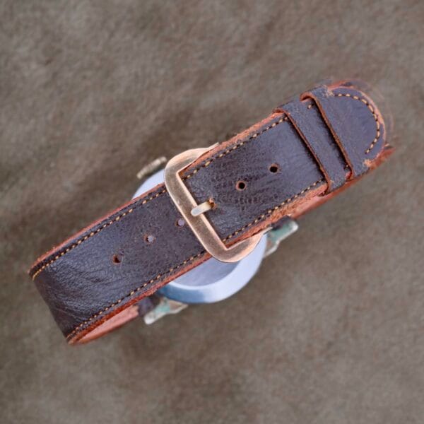 A brown leather strap with orange stitching and a silver buckle complements the attached watch, echoing the timeless elegance of Girard Perregaux Military Dial Small Seconds. The round silver case sits on a textured gray surface, reminiscent of classic military design.