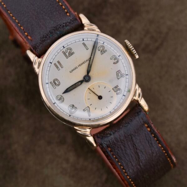 The Girard Perregaux Military Dial Small Seconds wristwatch features a round, aged white dial with Arabic numerals and black hands. It has a military-inspired small seconds subdial at 6 oclock, enclosed in a gold-toned case with a dark brown leather strap.