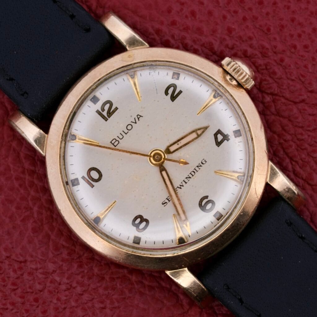 The Bulova Yankee Clipper Self-Winding watch boasts a vintage round gold-tone case and black leather strap. Its white dial features black numerals, the brand name, and Self-Winding text, all elegantly displayed on a textured red surface.