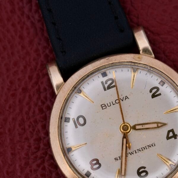 A close-up of the Bulova Yankee Clipper Self-Winding watch showcases a white face, gold hour markers, black leather strap, and hands poised at 10:10, elegantly resting on a textured burgundy surface.