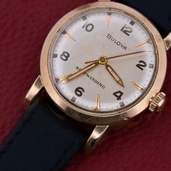 A vintage Bulova Yankee Clipper Self-Winding wristwatch with a gold case, white dial, black hour markers and numerals, leather strap, set against a textured reddish surface.