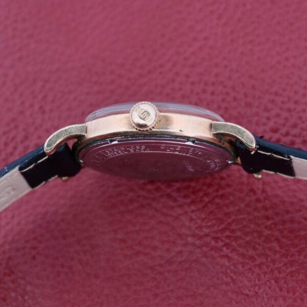 Side view of the Bulova Yankee Clipper Self-Winding watch featuring a gold case and crown on a dark leather strap. The gray back of the watch is visible against a textured burgundy background.