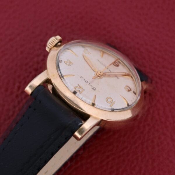 Displayed on a textured red surface, the Bulova Yankee Clipper Self-Winding wristwatch features a gold-toned case, black leather strap, cream-colored dial, and gold hour markers and hands.