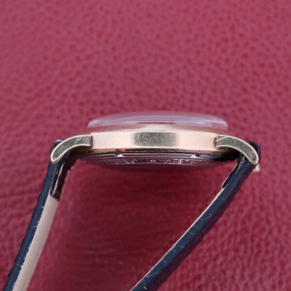 Side view of a Bulova Yankee Clipper Self-Winding gold watch with a black leather strap against a textured maroon background, highlighting its elevated face and slim profile.