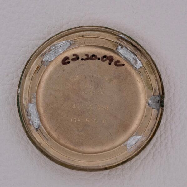 A close-up of the round metallic face of a Gruen Precision Cal. 510 Odd Lugs watch, featuring engraved text and a handwritten date at the top against a light textured background.