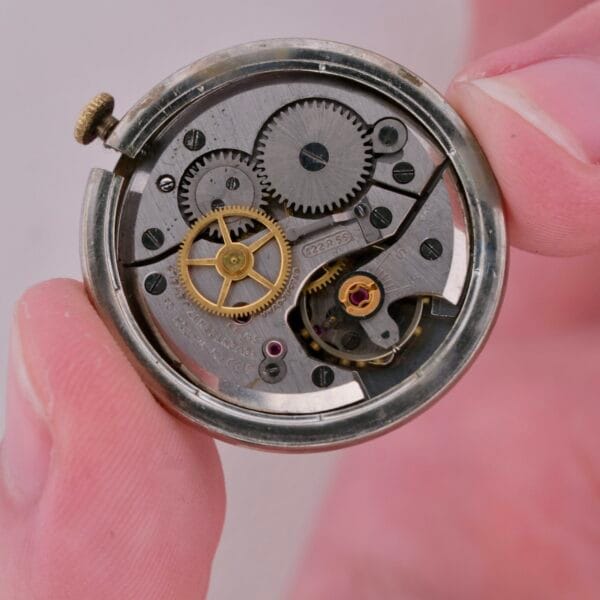 Close-up of a hand holding an open Gruen Precision Cal. 510 Odd Lugs watch movement, revealing intricate gears and a winding mechanism. The visible metal components and small wheels showcase the inner workings of this precise timepiece.