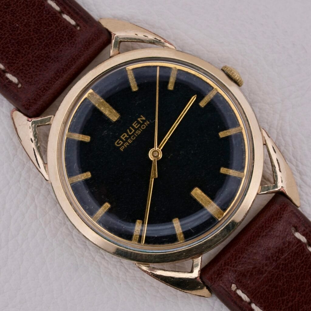 A vintage Gruen Precision Cal. 510 Odd Lugs wristwatch features a black dial with gold hour markers and hands, a gold-toned case, and a brown leather strap against a white background.