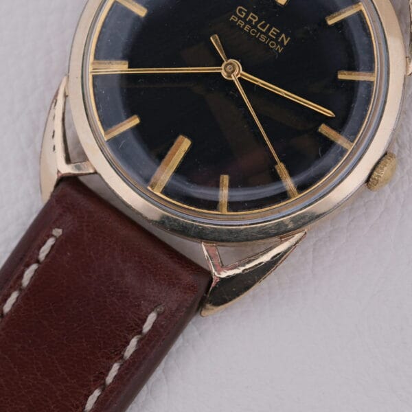 The Gruen Precision Cal. 510 Odd Lugs wristwatch features a black face, gold hour markers, and a brown leather strap with white stitching, elegantly displayed on a pristine white surface.