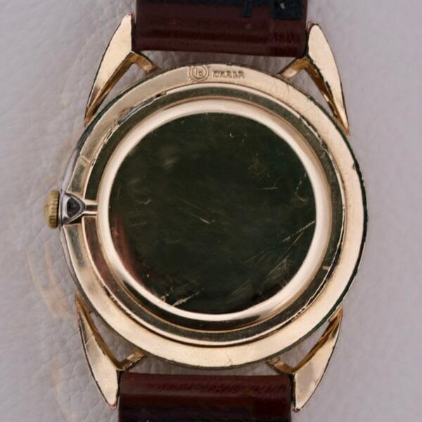 The image highlights the back of a vintage Gruen Precision Cal. 510 Odd Lugs wristwatch with a scratched gold-toned case. It features leather straps and is elegantly displayed on a textured light surface.