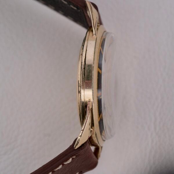 Side view of a vintage Gruen Precision Cal. 510 Odd Lugs wristwatch, featuring a gold-colored case and brown leather strap. The watch face is partially visible through a convex crystal against a white background, highlighting its intricate design.