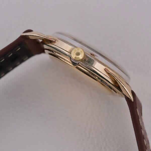A close-up of the Gruen Precision Cal. 510 Odd Lugs watch shows a gold case, brown leather strap, and crown on a white background. The watchs slight wear and tear enhance its timeless design.