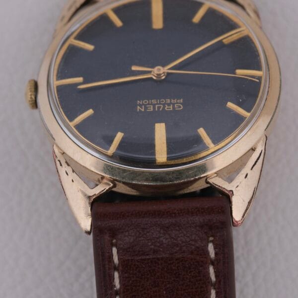 The Gruen Precision Cal. 510 Odd Lugs wristwatch features a dark face with gold markers and hands, complemented by a gold-tone case and brown leather strap, set against a textured white background.
