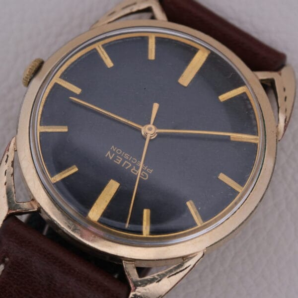 The Gruen Precision Cal. 510 Odd Lugs wristwatch features a dark face with gold hour markers and hands, a brown leather strap, and an elegant gold-toned metal case displaying Cal. 510 on its textured surface.