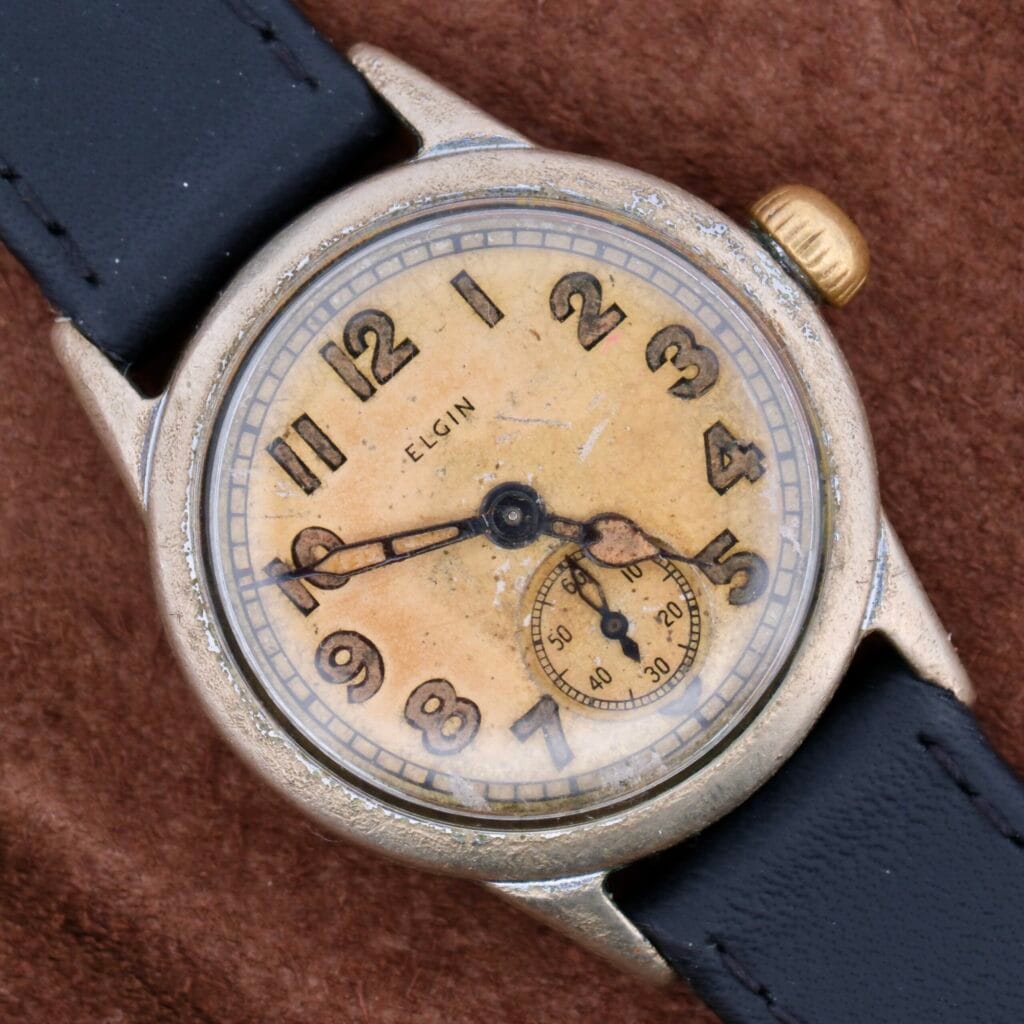 The WWII Military ELGIN General Service Watch features a worn cream dial with Arabic numerals, central hands, and a subdial at 6 oclock in a tarnished silver case. It comes with a black leather strap and rests on a brown surface.