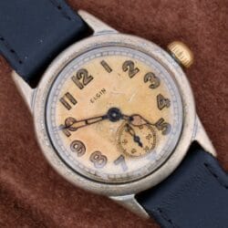 The WWII Military ELGIN General Service Watch features a worn cream dial with Arabic numerals, central hands, and a subdial at 6 oclock in a tarnished silver case. It comes with a black leather strap and rests on a brown surface.