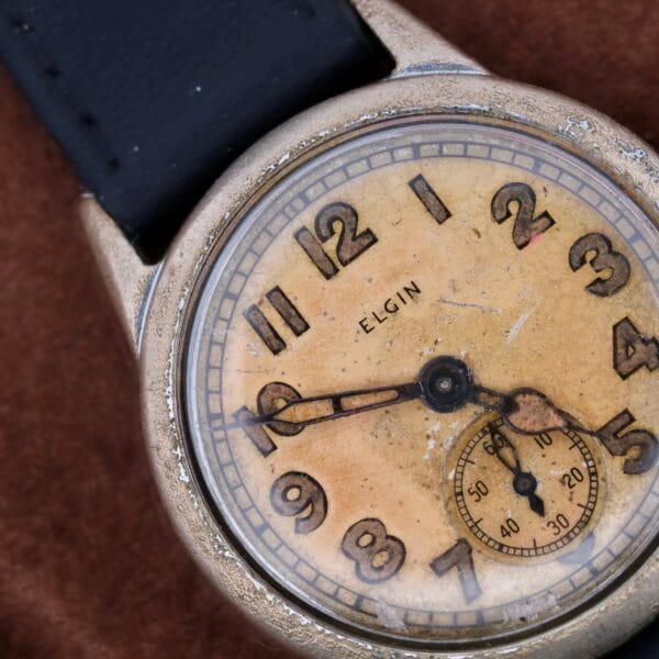 A close-up of a WWII Military ELGIN General Service Watch ORD. DEPT. U.S.A. 1944, showcasing its worn, yellowed dial with black numerals, a small seconds subdial at the bottom, and a black leather strap. The watchcase displays signs of wear and aging.