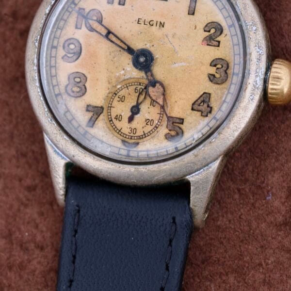 Close-up of a WWII Military ELGIN General Service Watch ORD. DEPT. U.S.A. 1944 with a weathered dial, featuring bold black numerals and a small seconds dial at the bottom. Its black leather strap rests on a textured brown surface, exuding timeless charm from its storied past.
.