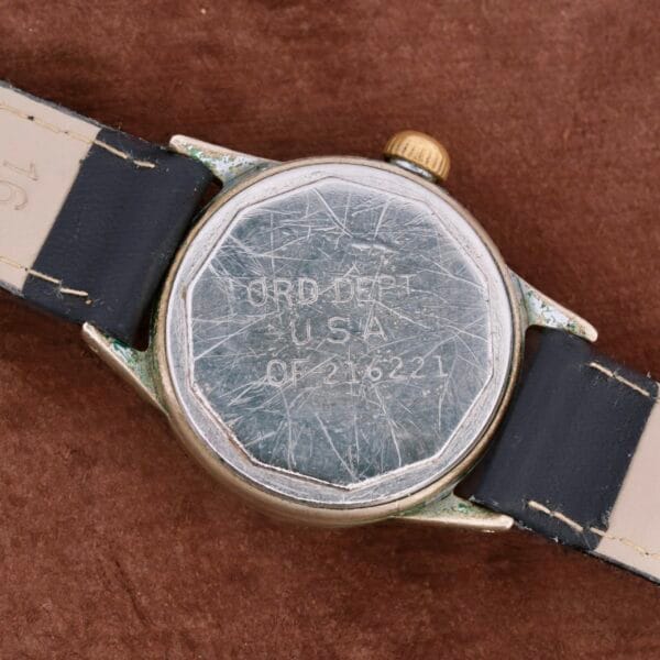 Back view of a weathered WWII Military ELGIN General Service Watch with black and white strap, displaying scratches on the metal case and engraved ORD. DEPT. U.S.A. OF216221, set on a brown textured surface.