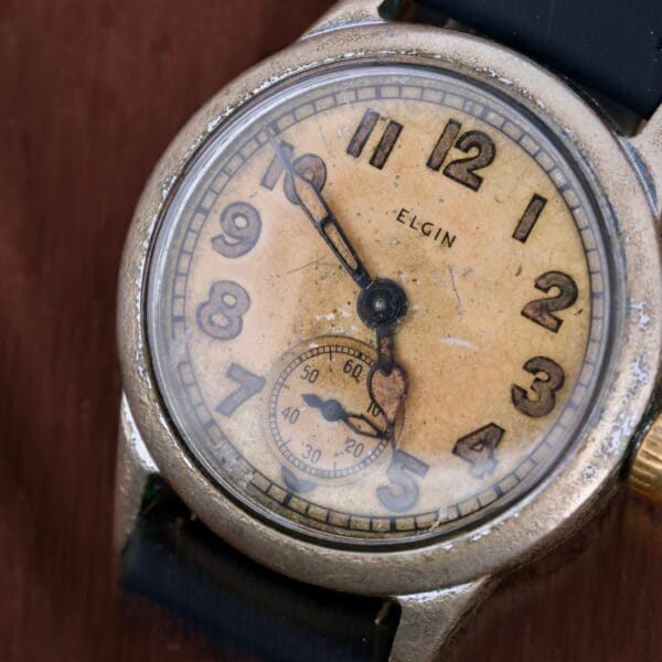 Close-up of the vintage WWII Military ELGIN General Service Watch ORD. DEPT. U.S.A. 1944, featuring a weathered dial with bold numbers and a smaller seconds subdial, all mounted on a dark strap that enhances its antique charm and storied past.