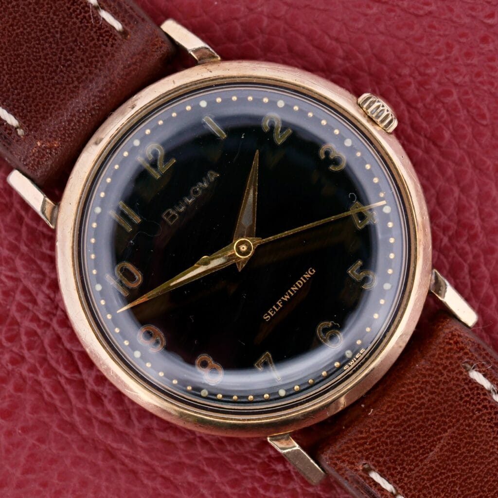 The vintage Bulova Automatic Black Dial Arabic Numerals Watch features a self-winding mechanism, a black dial with gold numerals and hands, gold casing, and a brown leather strap, elegantly displayed against a textured red surface.