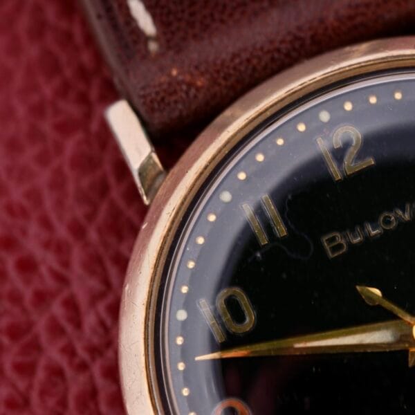 Close-up of a vintage Bulova Automatic Black Dial Arabic Numerals watch with gold markers, set in a gold case and paired with a brown leather strap. Minute and hour hands indicate ten and two against a textured red backdrop.
