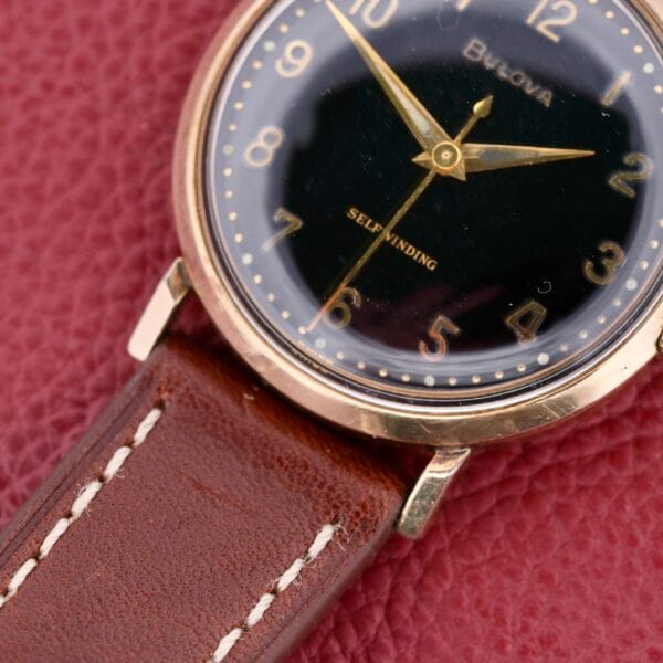 Close-up of a vintage Bulova Automatic Black Dial Arabic Numerals watch, featuring a gold case and brown leather strap with white stitching, set against a textured red background.