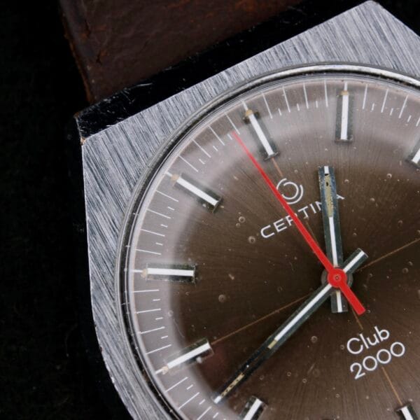 Close-up of a 1970s Certina Club 2000 Cal. 2502 watch with a silver bezel, brown dial, and red second hand. The white minute and hour hands have luminous markers, and the brand and model name are clearly visible on the dial.