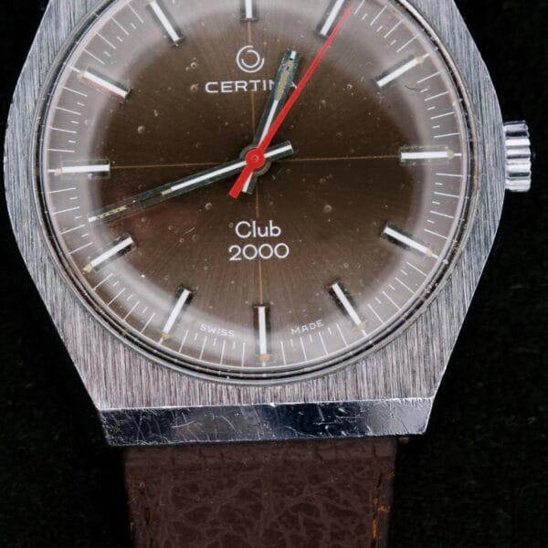 The Certina Club 2000 Cal. 2502 is a vintage 1970s watch featuring a silver brushed metal case, brown dial, and striking red second hand. It includes silver hour markers with Swiss Made at the bottom and is paired with a textured dark brown leather strap.