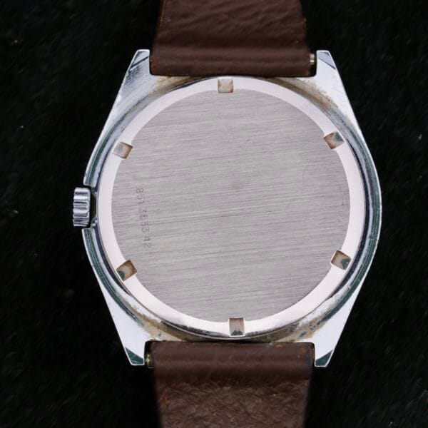 The image features the back of a 1970s Certina Club 2000 Cal. 2502 wristwatch, showcasing a round metal case with a smooth surface and minimal engravings. This vintage piece is enhanced by a brown leather strap attached to its polished silver frame.