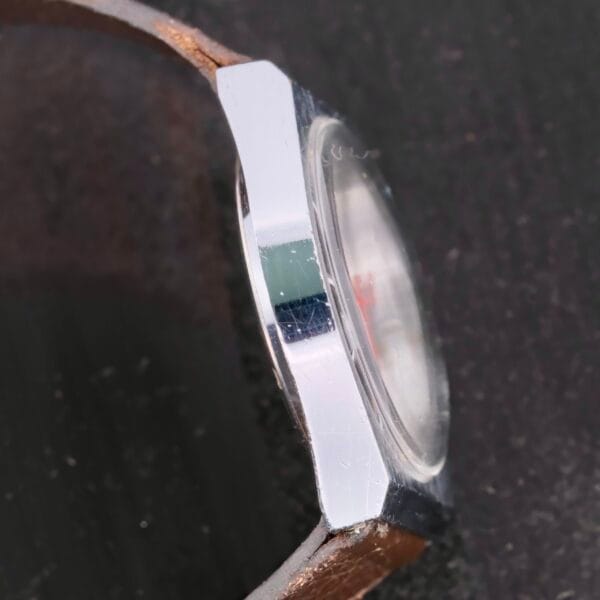 Side view of a Certina Club 2000 Cal. 2502 wristwatch with a metallic case and brown leather strap, highlighting its shiny, thick edge against a dark, textured background.